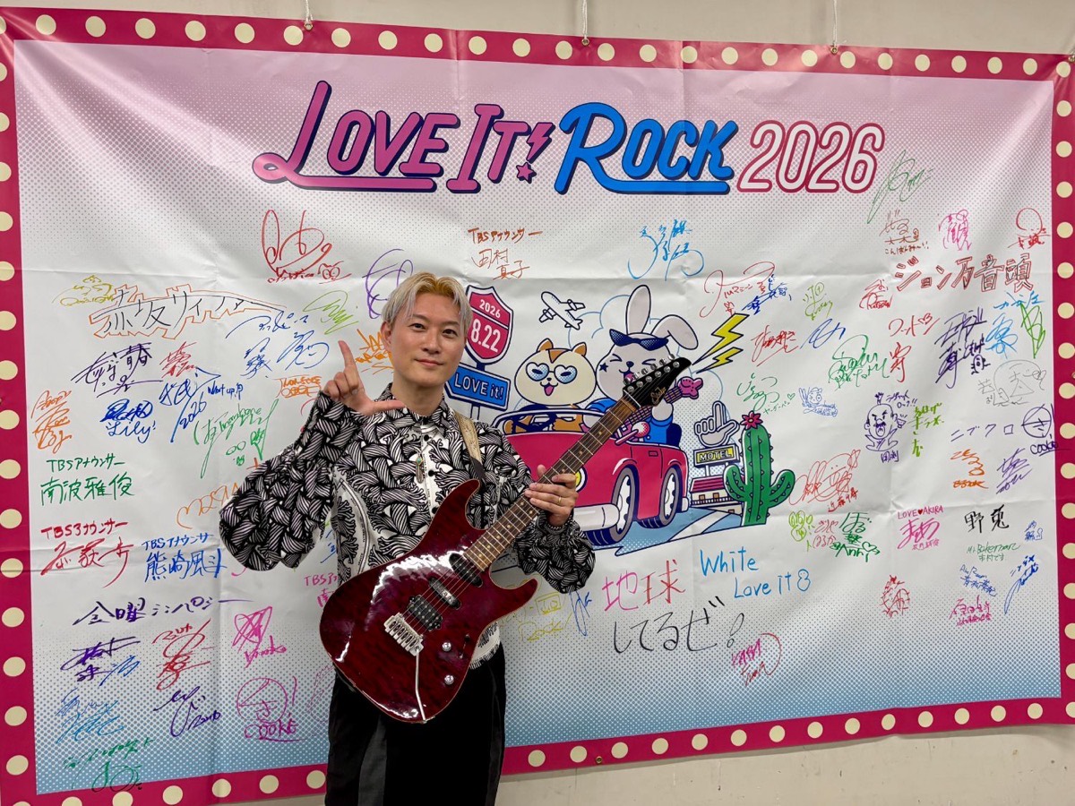 ラヴィット！ロック2026 出演者サイン入りボードの前でギターを持つ講師Kaz Takayama